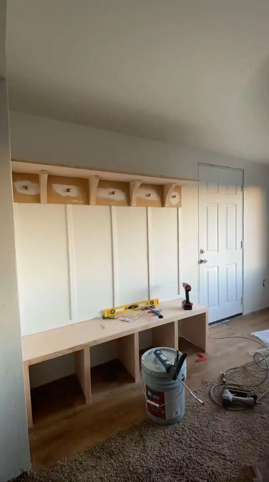 A nearly finished built-in entryway bench and shelf with board and batten detailing. The wooden storage unit is installed against a white wall, with tools and a bucket of construction adhesive still present as the final touches are completed.